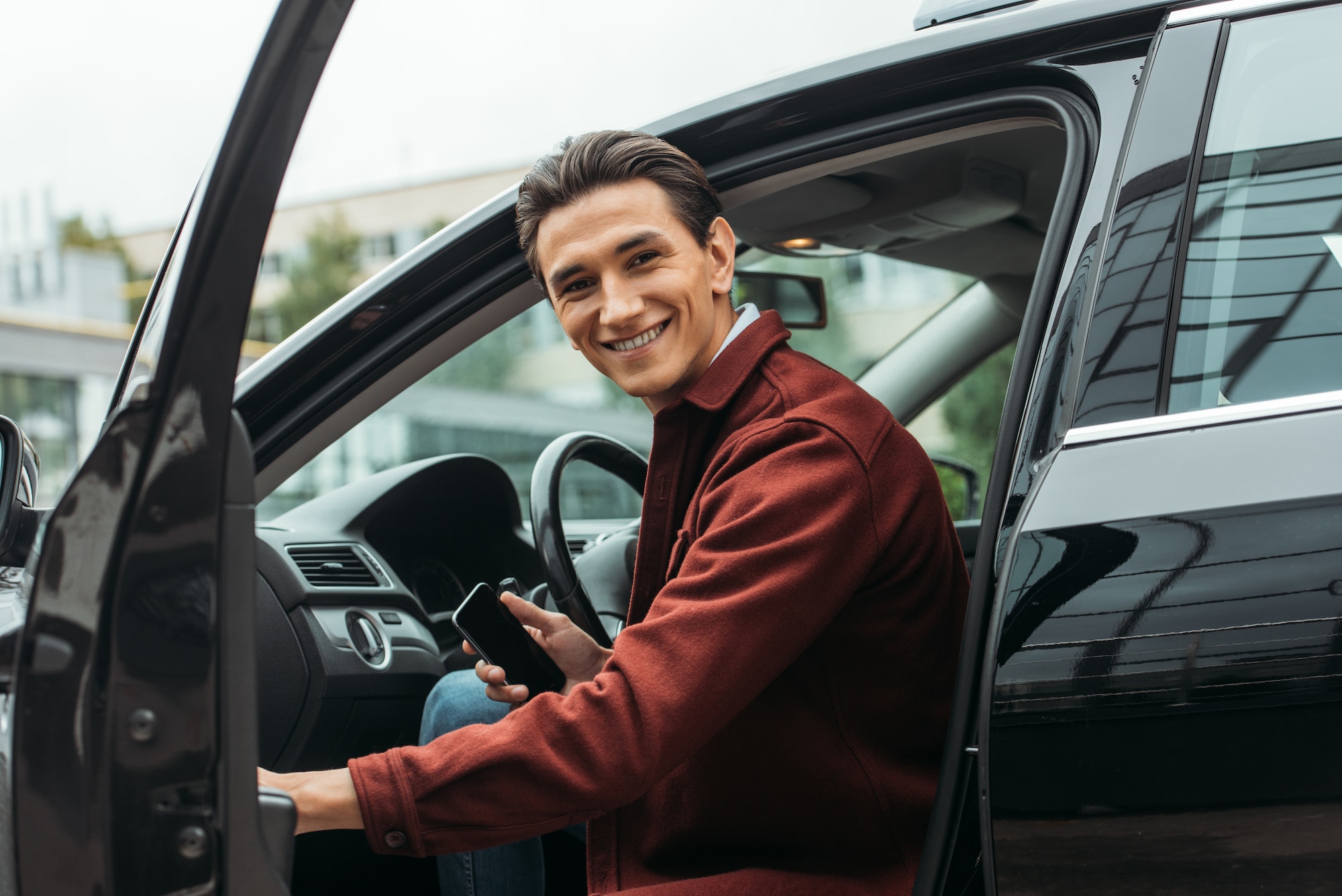 Smiling Taxi Driver Sitting In Car With Open Door And Holding Smartphone